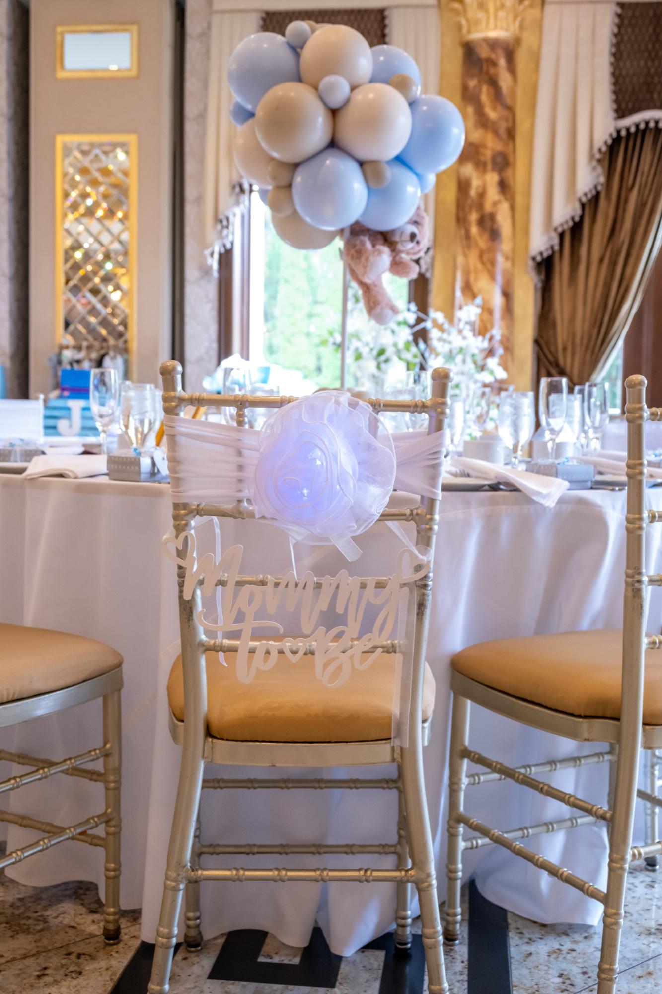 Decorated chair, table settings, and blue balloons prepared for a baby shower.