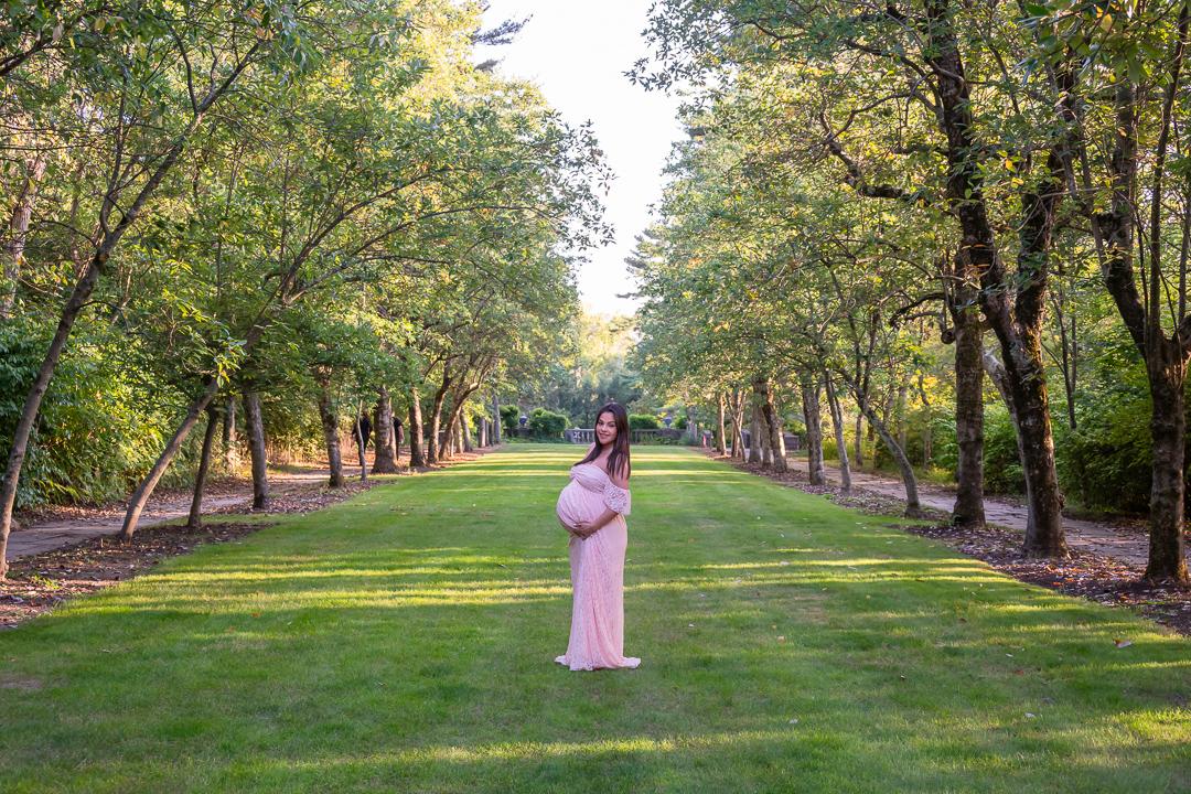 Expectant mother in a pale pink dress standing in a tree-lined garden.